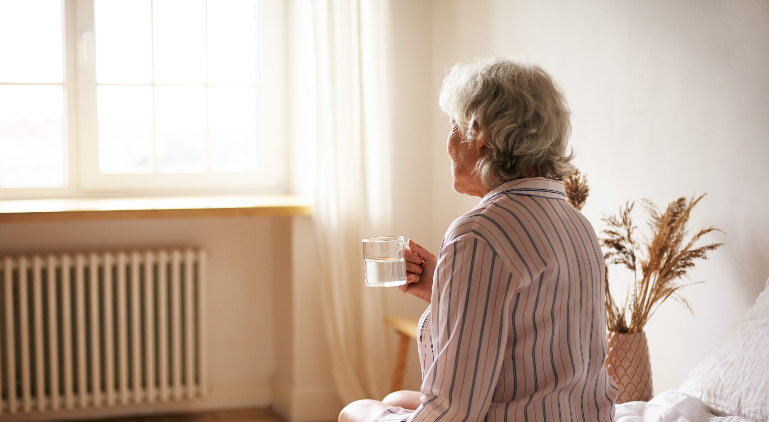 Rear view of senior sixty year old woman with gray hair holding mug washing down sleeping pill, suffering from insomnia. Elderly retired female taking medicine with water, sitting in bedroom